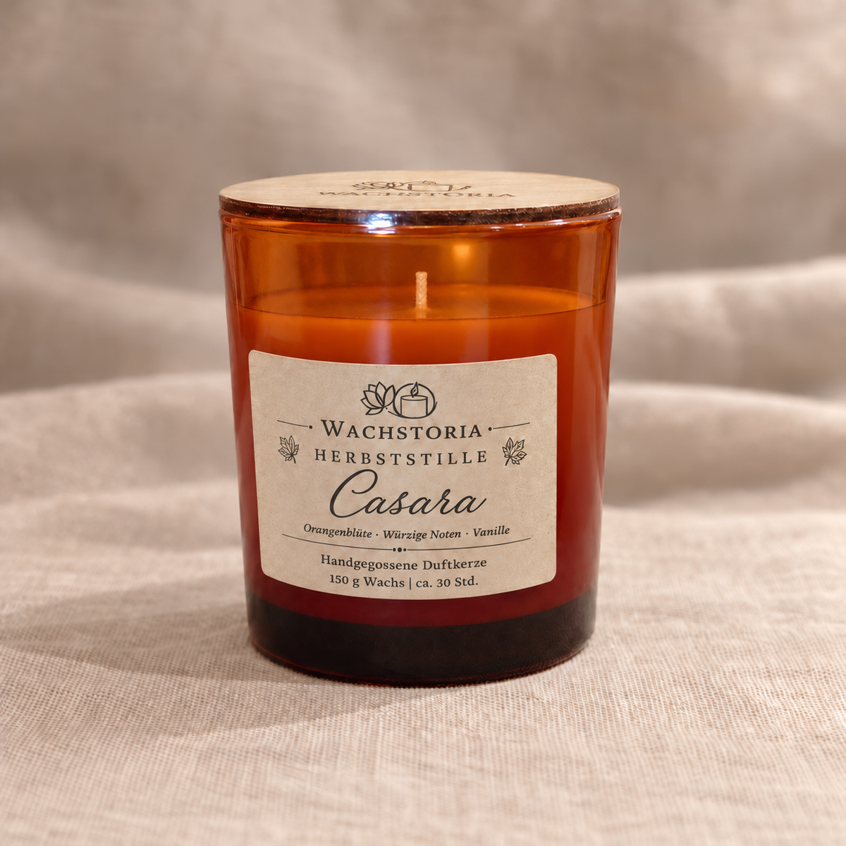 Wachstoria Duftkerze Casara, 150 g, im bernsteinfarbenen Glas mit dünnem Holzdeckel und Kraftpapieretikett auf beigem Leinen.