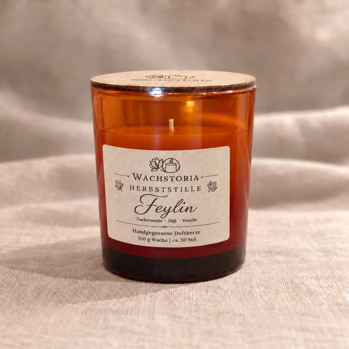 Wachstoria Duftkerze Feylin, 150 g, im bernsteinfarbenen Glas mit dünnem Holzdeckel und Kraftpapieretikett auf beigem Leinen.