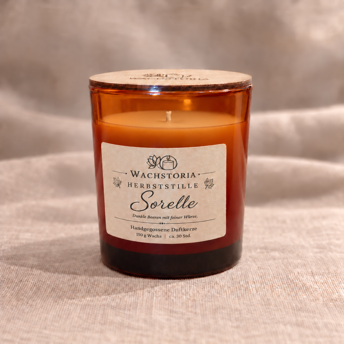 Wachstoria Duftkerze Sorelle, 150 g, im bernsteinfarbenen Glas mit dünnem Holzdeckel und Kraftpapieretikett auf beigem Leinen.