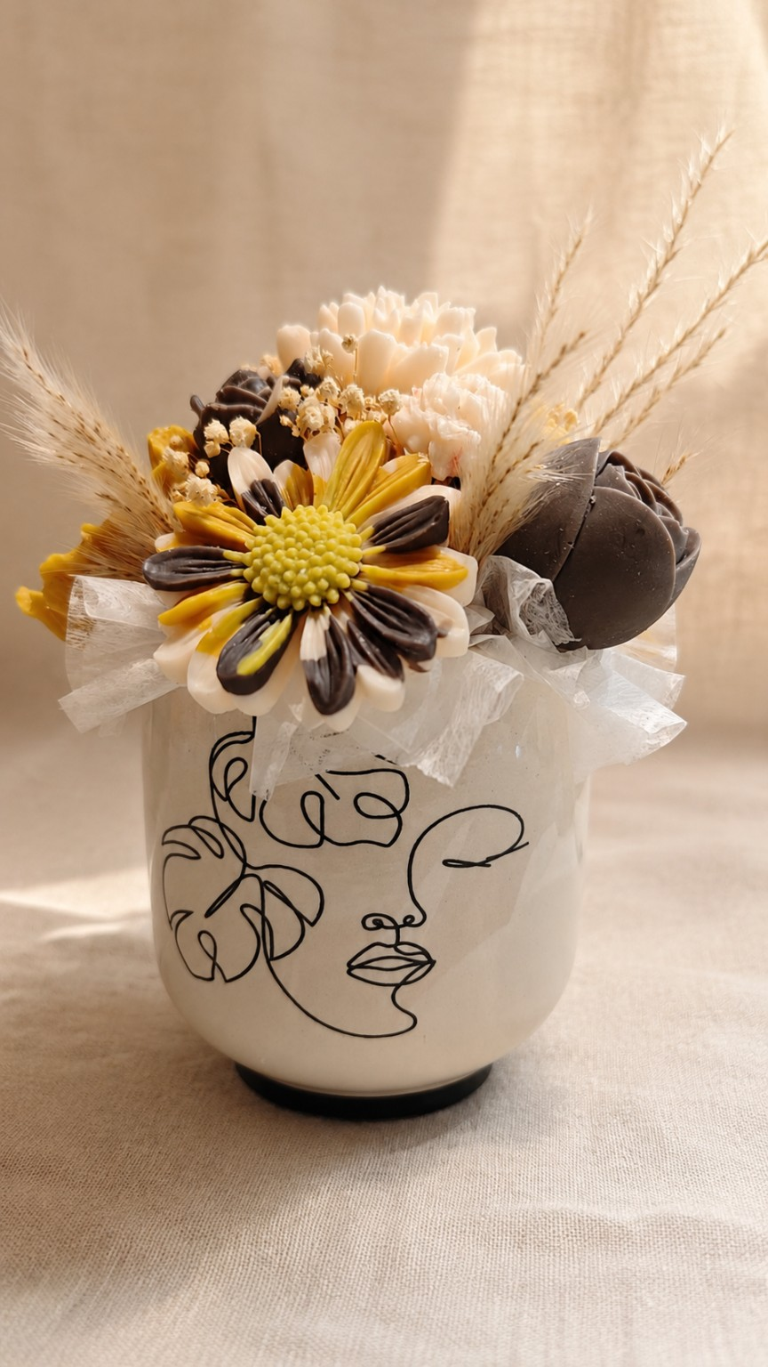 Wachstoria Wachsblumenstrauß mit gelben, cremefarbenen und braunen Wachsblüten, Trockenblumen und einer Vase mit schwarzer Gesichtszeichnung.
